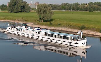A white boat named Fluvius travels along a river, surrounded by green fields and trees under a&hellip;