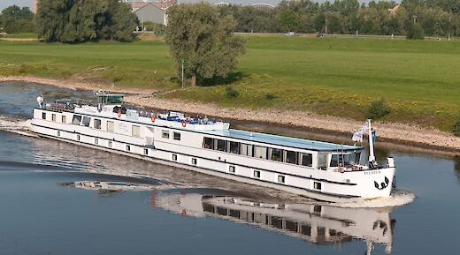 A white boat named Fluvius travels along a river, surrounded by green fields and trees under a clear blue sky.