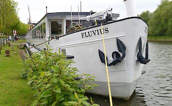 The front view of a white boat named Fluvius docked beside a grassy bank, with ropes securing it to&hellip;