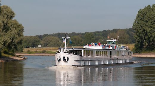 A white boat named Fluvius moves smoothly along a river, surrounded by green trees and grasslands under a clear sky.