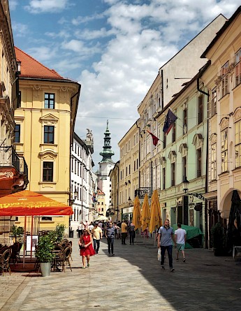 St. Michael's Gate in Bratislava, Slovakia, with people walking along a historic street lined with colorful buildings and outdoor cafés under a partly cloudy sky.