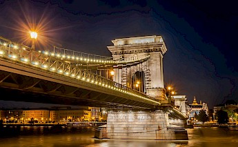 A nighttime view of the illuminated Chain Bridge in Budapest, with city lights reflecting on the Danube River.