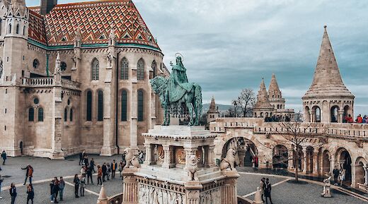 The Fisherman's Bastion in Budapest, featuring ornate architecture with pointed towers and a statue of a rider on horseback. People are walking around the square under a cloudy sky.