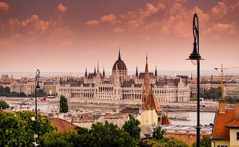 The Hungarian Parliament Building in Budapest along the Danube River, under a cloudy sky, with street lamps and rooftops in the foreground.