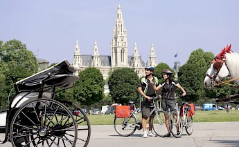 A man and a woman with bicycles stand in front of a horse-drawn carriage, with a large cathedral visible in the background.