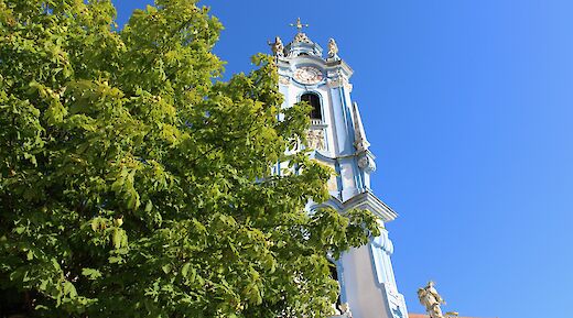 A view of the blue and white bell tower of the D&uuml;rnstein Abbey in Austria, partially obscured by lush green trees against a clear blue sky.