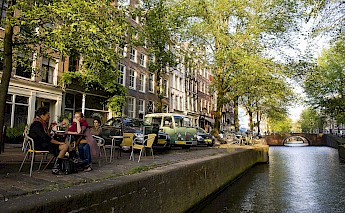 People sitting outdoors at a café along a canal in Amsterdam with traditional buildings and trees lining the street.