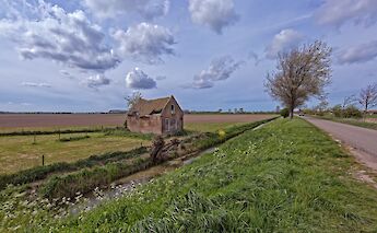 A rural scene in the Dutch countryside featuring an old, weathered house by a canal with expansive fields under a partly cloudy sky.