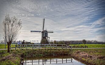 Cyclists riding on a path beside a traditional Dutch windmill in a rural setting with a canal in the foreground.