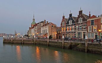 A waterfront scene in Dordrecht, Netherlands, with historic gabled buildings reflected in the water at dusk.