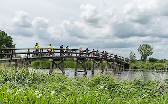 Cyclists riding across a wooden bridge over a river, surrounded by greenery and cloudy sky.