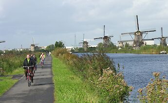Cyclists riding along a path beside a river with windmills in the background in Kinderdijk, Holland.