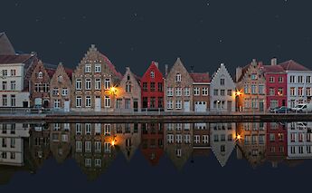 Row of narrow, traditional Flemish buildings along a canal in Bruges, Belgium, reflected in the water at night.
