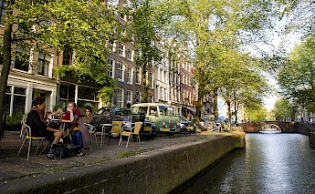 A canal in Amsterdam, lined with bicycles and a tree-shaded cafe where people sit, with traditional Dutch buildings and a bridge visible in the background.