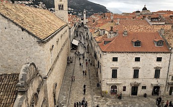 An aerial view of Dubrovnik’s historic center, showing a bustling street lined with buildings that have distinctive red-tiled roofs and stone walls.
