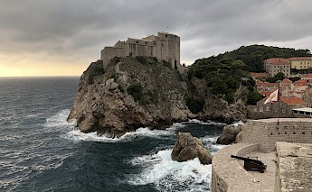 A fortress perched on a rocky cliff overlooking the sea in Dubrovnik, Croatia, with waves crashing against the rocks under a cloudy sky.