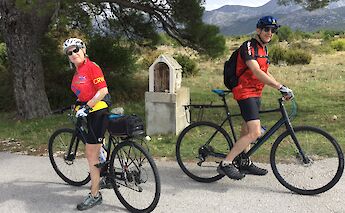 Two cyclists in colorful sports attire taking a break on a rural road in Croatia, surrounded by trees and rocky terrain.