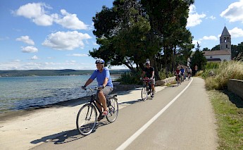 Cyclists on a path alongside water, with trees and a building with a tower in the background, in Dalmatia, Croatia.