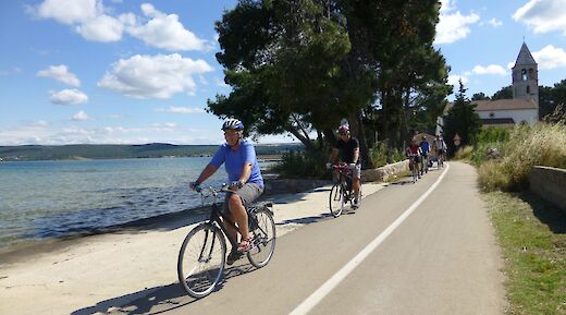 Cyclists on a path alongside water, with trees and a building with a tower in the background, in Dalmatia, Croatia.