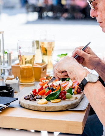 A person dining outdoors in Croatia with a wooden platter of assorted fruits and vegetables, and glasses of beer on the table.