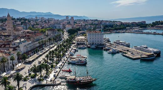 Aerial view of a coastal town in Split, Dalmatia, Croatia, featuring a promenade lined with palm trees, boats docked at the harbor, and surrounding buildings.
