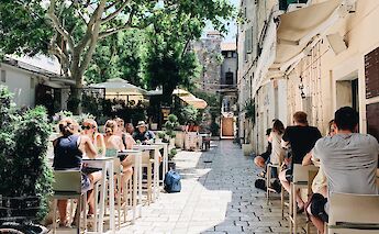 People seated at outdoor tables in a sunny, narrow alley surrounded by trees and historic buildings&hellip;