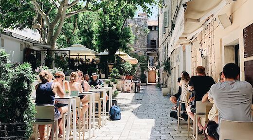 People seated at outdoor tables in a sunny, narrow alley surrounded by trees and historic buildings in Split, Croatia.