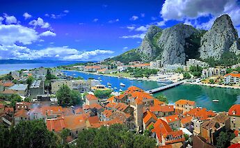 A panoramic view of a coastal town in Split, Dalmatia, Croatia, with red-roofed buildings, a winding river, and dramatic rocky cliffs under a bright blue sky.