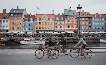 Cyclists ride along the waterfront in Nyhavn, Copenhagen, with colorful buildings and boats in the background.