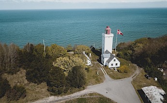 The image shows a lighthouse and several buildings near the coast surrounded by trees, with a Danish flag flying on the property in Gilleleje, Denmark. The sea is visible in the background.