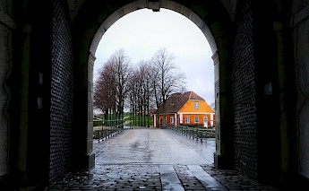 View from under an archway leading to a cobblestone path with a bright orange building in Helsingør, Denmark. Bare trees line the path on a wet day.