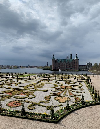 Frederiksborg Castle in Hillerød, Denmark, with an elaborate garden in the foreground under a cloudy sky.