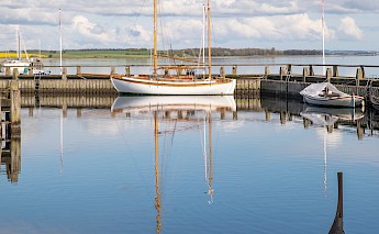A wooden sailboat and other small boats docked at a calm harbor in Roskilde, Denmark, under a partly cloudy sky.
