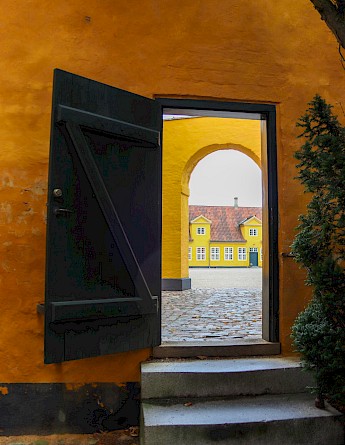 A wooden door partially open to reveal a cobblestone path leading to a colorful yellow building with red roof tiles in Roskilde, Denmark.
