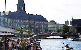 A busy canal scene in Copenhagen with people enjoying outdoor seating by the water. There are large historic buildings, a bridge, and boats on the canal.