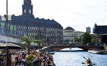 A lively canal scene in Copenhagen with people gathered near the water, boats in the canal, and historic buildings and a bridge in the background.