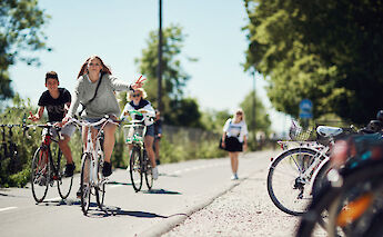 People cycling on a path lined with greenery, likely in Denmark. A young woman in the foreground is gesturing with a peace sign while riding.