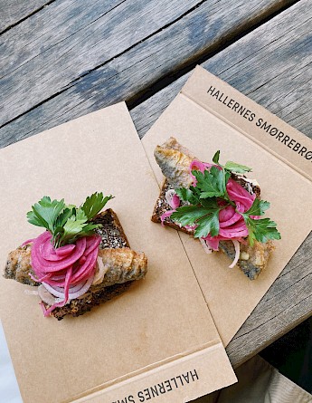 Two pieces of smørrebrød topped with fish, pickled onions, and parsley on a wooden table.