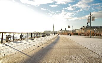 A waterfront promenade in Bordeaux, France, with historic buildings and a spire visible in the background.