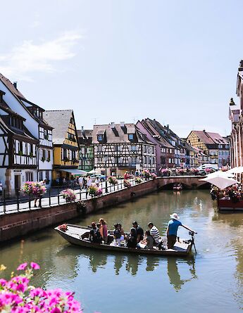 A boat with passengers on a canal in Colmar, France, surrounded by colorful, traditional half-timbered houses and flowers under a clear sky.