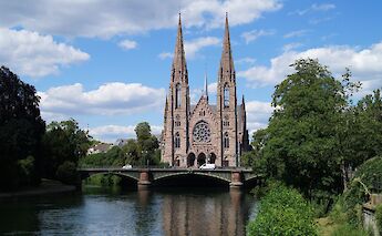 A cathedral with twin spires by a river, set against a partly cloudy sky in Strasbourg, France.