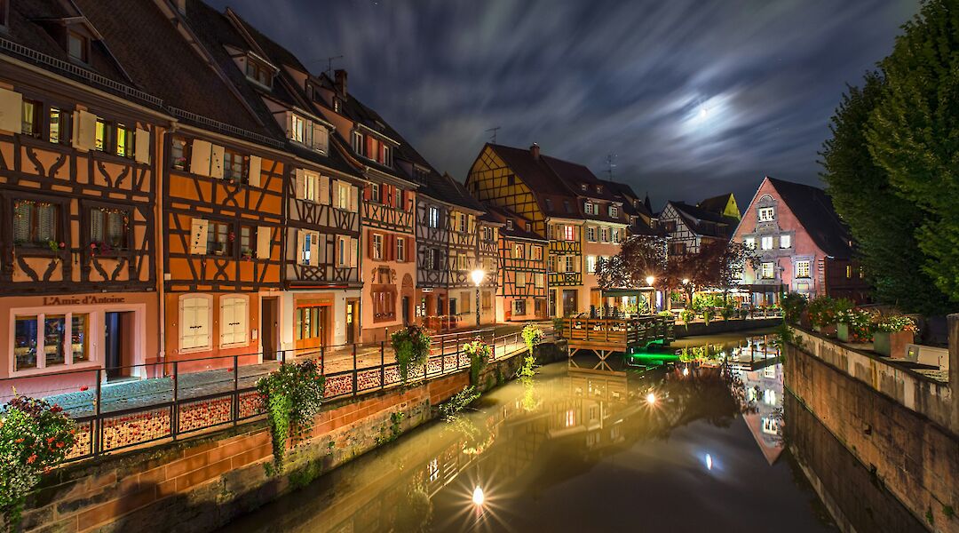 Timber-framed buildings reflecting in a canal at night in Colmar, France, with the moon illuminating the sky.