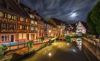 Timber-framed buildings reflecting in a canal at night in Colmar, France, with the moon illuminating the sky.