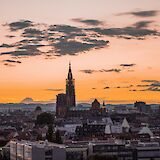 The Strasbourg skyline at sunset, featuring the cathedral spire silhouetted against an orange sky, with scattered clouds above the cityscape.