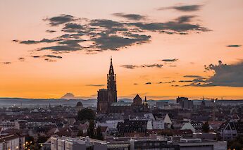 The Strasbourg skyline at sunset, featuring the cathedral spire silhouetted against an orange sky, with scattered clouds above the cityscape.
