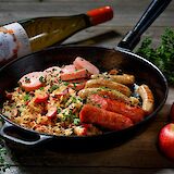 Sausages and sauerkraut in a pan, accompanied by apples and a bottle of wine, on a rustic wooden table.