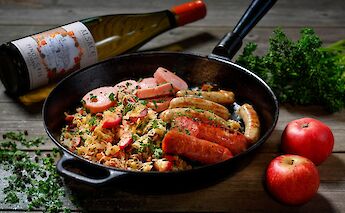 Sausages and sauerkraut in a pan, accompanied by apples and a bottle of wine, on a rustic wooden table.