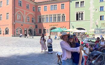 People walking and sitting near colorful historical buildings in a square in Regensburg, Germany.