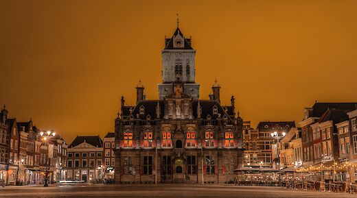 A historic building with a clock tower in an illuminated square with a warm orange sky.