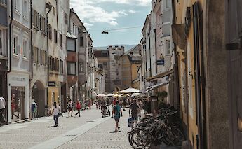 A bustling street in an Italian town with people walking and cycling, lined with colorful buildings and outdoor cafés.